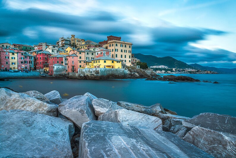 May include: A colorful Italian village with pastel-colored buildings sits on a rocky coastline. The water is a deep blue and the sky is a light blue with white clouds. The village is surrounded by lush green hills.