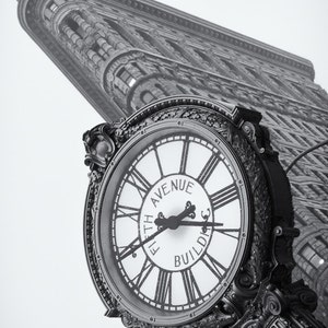 Puede incluir: Una fotografía en blanco y negro de una esfera de reloj con números romanos y las palabras "Flatiron Avenue Building". El reloj está unido a una estructura metálica decorativa. El Flatiron Building está en el fondo.