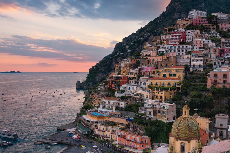 May include: A colorful town built on a steep cliffside overlooking the ocean at sunset. The town is filled with buildings of various sizes and colors, with a large, green-domed church at the bottom of the image.