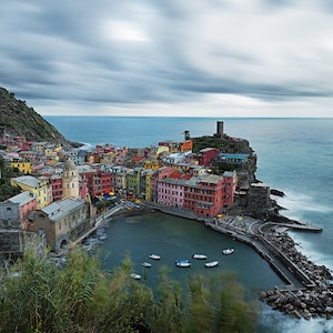 May include: A picturesque Italian village nestled on a cliffside overlooking the turquoise sea. Colorful buildings line the harbor, with boats bobbing in the water. The sky is overcast with dramatic clouds.