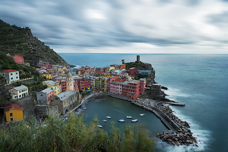 May include: A picturesque Italian village nestled on a cliffside overlooking the turquoise sea. Colorful buildings line the harbor, with boats bobbing in the water. The sky is overcast with dramatic clouds.