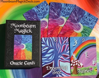 May include: A deck of "Moonbeam Magick" oracle cards with a black box and several cards fanned out. The box and cards feature colorful artwork with a crescent moon and rainbow hues. Two cards show the words "Dreaming Tree" and "Fairies".