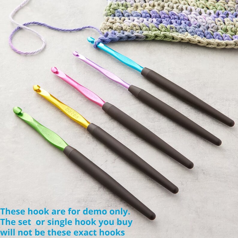 May include: A set of five crochet hooks with colorful metal heads and dark gray ergonomic handles. The hooks are in shades of green, gold, pink, purple, and blue. The text on the hooks reads "Bates". A crocheted swatch and yarn are in the background.