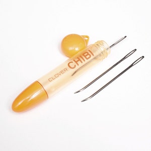 May include: A clear plastic container with a yellow cap and the text "CLOVER CHIBI" on the side. The container holds two silver needles.