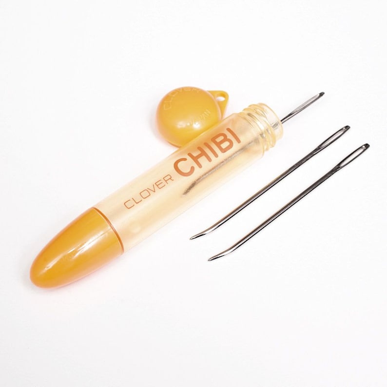 May include: A clear plastic container with a yellow cap and the text "CLOVER CHIBI" on the side. The container holds two silver needles.