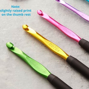 May include: Close-up of several colorful crochet hooks. The hooks are in shades of green, gold, pink, and purple. Each hook has a black handle and the text "Susan Bates" is visible on some. The text "Note: slightly raised print on the thumb rest" is also visible.