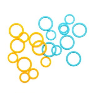 May include: A collection of yellow and blue circular stitch markers of varying sizes, scattered on a white background. These small, ring-shaped markers are used in knitting and crochet projects to indicate specific points in the pattern.