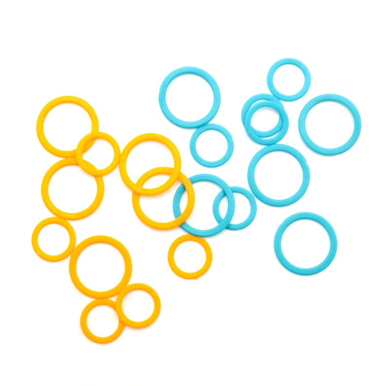 May include: A collection of yellow and blue circular stitch markers of varying sizes, scattered on a white background. These small, ring-shaped markers are used in knitting and crochet projects to indicate specific points in the pattern.