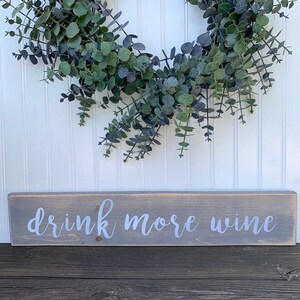 May include: A gray wooden sign with the text "drink more wine" written in white paint. The sign is hanging on a white wall with a green eucalyptus wreath above it.