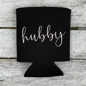 Puede incluir: Enfriador de latas de neopreno negro con la palabra "hubby" escrita en letra cursiva blanca.