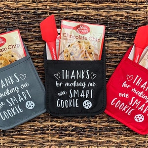 May include: Three oven mitts with pockets, each containing a bag of chocolate chip cookie mix and a red silicone spatula. The mitts are gray, black, and red, and each has the text "THANKS for making me one SMART COOKIE" printed on the front.