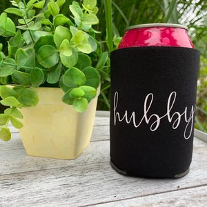 Puede incluir: Enfriador de latas de neopreno negro con la palabra "hubby" escrita en cursiva blanca.