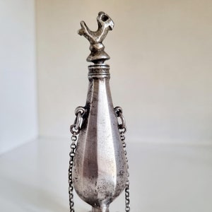 Peut inclure: Une bouteille en argent ornée d'un bouchon en forme d'oiseau et d'une chaîne attachée au col. La bouteille a une forme de larme et une base détaillée.