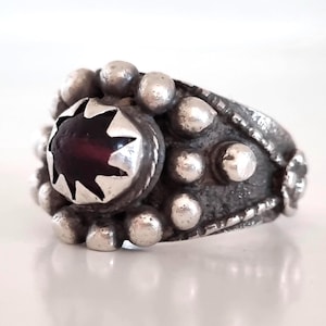 Può includere: Anello in argento con una pietra rosso scuro a forma di stella al centro. L'anello è circondato da piccole perline rotonde d'argento e ha una fascia strutturata. Design vintage.