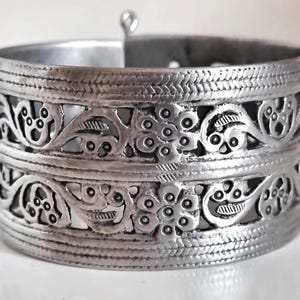 May include: A wide, silver cuff bracelet with detailed floral and leaf cut-out patterns. The metal band has textured detailing, giving a vintage appearance. The bracelet features a clasp for easy wear. The circumference is approximately 18 cm.