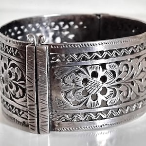 Könnte beinhalten: Ein silberfarbenes Armreif-Armband mit aufwendigen floralen und geometrischen Mustern. Das breite Band weist detaillierte Schnitzereien von Blumen und Blättern auf, mit einem sicheren Verschluss. Das Armband ist etwa 5 cm breit.