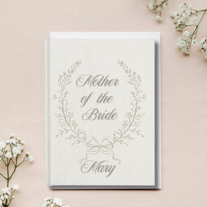 May include: A cream-coloured card featuring the text "Mother of the Bride" within a floral wreath, with the name "Mary" below. The card rests on a light pink surface, with white flowers in the background.