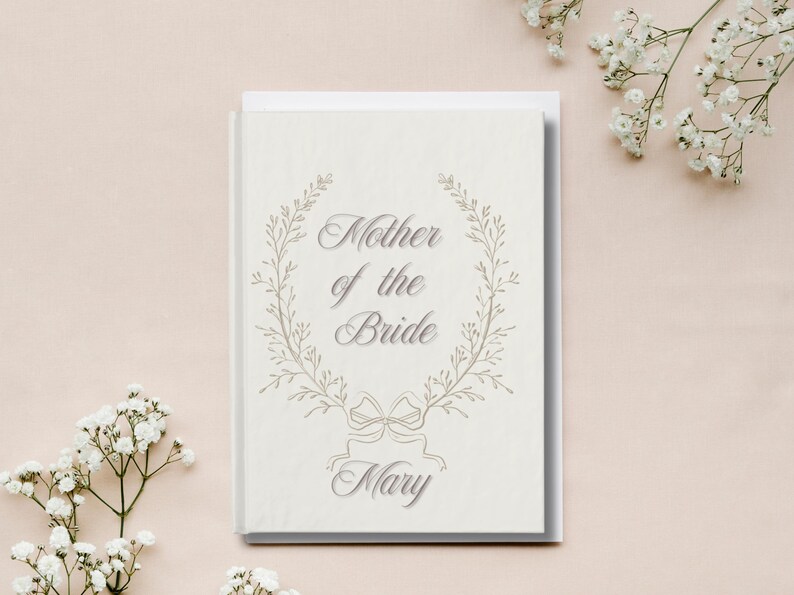 May include: A cream-coloured card featuring the text "Mother of the Bride" within a floral wreath, with the name "Mary" below. The card rests on a light pink surface, with white flowers in the background.