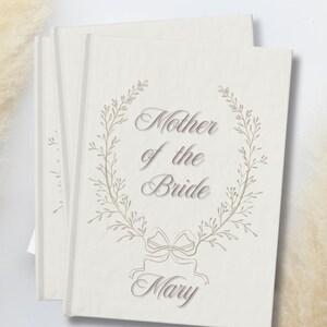 May include: A stack of cream-coloured cards featuring the text "Mother of the Bride" in an elegant script, surrounded by a delicate floral wreath. The name "Mary" is below. The cards have a soft, textured appearance.
