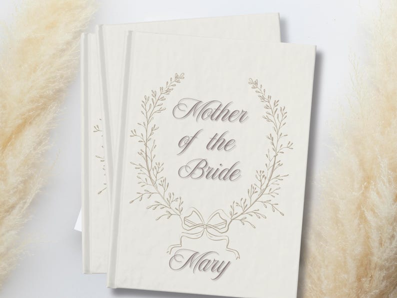 May include: A stack of cream-coloured cards featuring the text "Mother of the Bride" in an elegant script, surrounded by a delicate floral wreath. The name "Mary" is below. The cards have a soft, textured appearance.