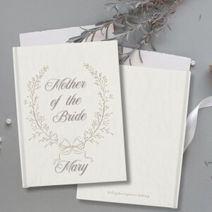 May include: Cream-coloured wedding guest book with the inscription "Mother of the Bride" and the name "Mary" in a decorative font, framed by a floral wreath. A second blank book and a spool of white ribbon are also visible.