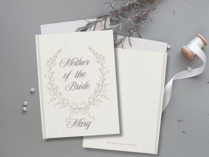 May include: Cream-coloured wedding guest book with the inscription "Mother of the Bride" and the name "Mary" in a decorative font, framed by a floral wreath. A second blank book and a spool of white ribbon are also visible.