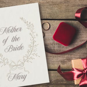 May include: A wedding-themed image featuring a white card with the text "Mother of the Bride Mary" surrounded by a floral wreath. A red ring box, two gold rings, and a gift box with a red ribbon on a wooden surface.
