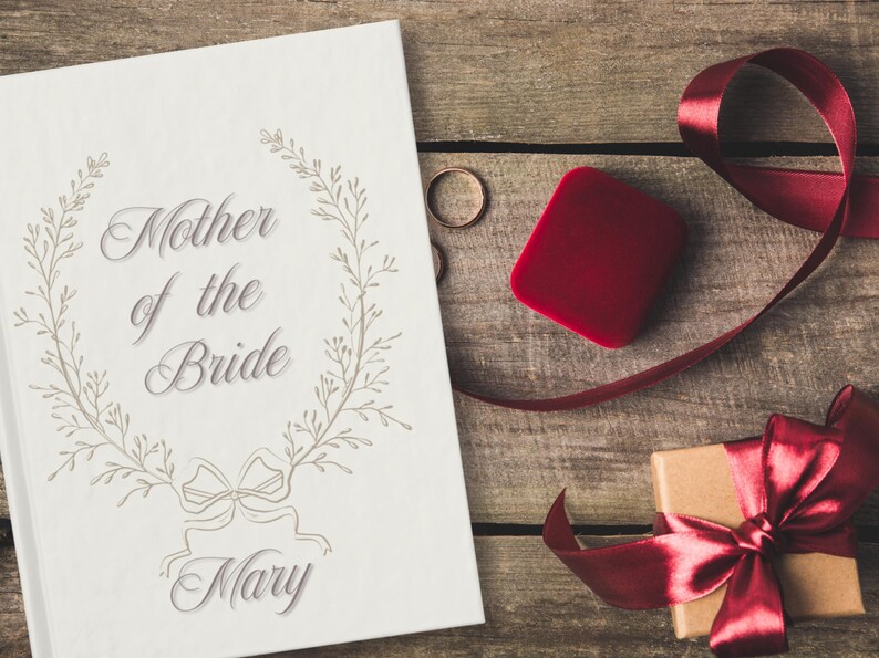 May include: A wedding-themed image featuring a white card with the text "Mother of the Bride Mary" surrounded by a floral wreath. A red ring box, two gold rings, and a gift box with a red ribbon on a wooden surface.