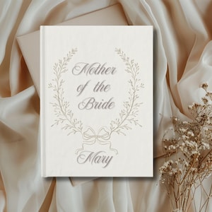 May include: A cream-coloured book featuring the words "Mother of the Bride" in a decorative font, encircled by a floral wreath. The name "Mary" is displayed below. The book rests on a backdrop of flowing fabric and dried flowers.