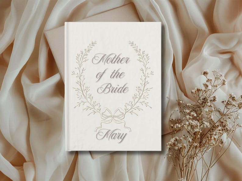 May include: A cream-coloured book featuring the words "Mother of the Bride" in a decorative font, encircled by a floral wreath. The name "Mary" is displayed below. The book rests on a backdrop of flowing fabric and dried flowers.