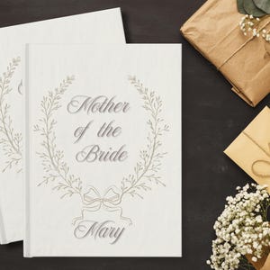 May include: Two white hardcover books with the text "Mother of the Bride" and the name "Mary" in elegant script, surrounded by a floral wreath design. The image also includes wrapped gifts and a card.
