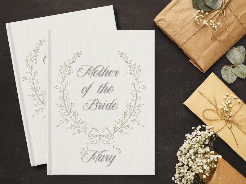 May include: Two white hardcover books with the text "Mother of the Bride" and the name "Mary" in elegant script, surrounded by a floral wreath design. The image also includes wrapped gifts and a card.