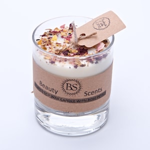 Peut inclure: Une bougie en verre transparent avec une base de cire blanche et une étiquette en papier brun qui indique "Beauty Scents" et "Handcrafted with Love". La bougie est décorée de pétales de rose séchés et d'autres éléments botaniques.