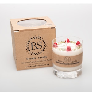 May include: A white soy wax candle with a raspberry scent. The candle is in a clear glass jar with a brown label that reads "Beauty Scents Scented Soy Wax Candle with Raspberry". The candle is topped with red raspberries and green herbs.