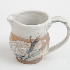 May include: A small, white ceramic pitcher with a brown and blue floral design. The pitcher has a rounded body and a handle.