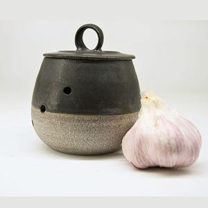 May include: A black and grey ceramic garlic keeper with a lid and a small handle. The keeper has a textured, speckled grey base and a smooth black top. A bulb of garlic rests beside the keeper.