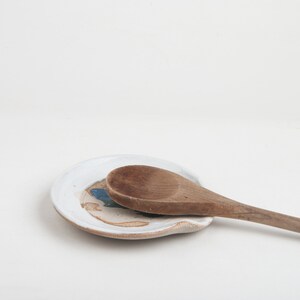 Peut inclure: Un repose-cuillère en céramique blanche avec un motif bleu et brun. Une cuillère en bois repose sur le repose-cuillère.
