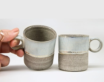 Pode incluir: Duas canecas de cerâmica feitas à mão. As canecas têm uma metade superior esmaltada em azul claro e uma metade inferior texturizada em cinza. Uma fina faixa dourada separa as duas seções. Cada caneca tem uma alça pequena.