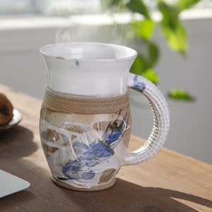 May include: A white ceramic mug with a brown and blue design. The mug has a unique, textured handle and is filled with a steaming beverage.