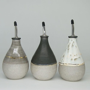 Peut inclure: Trois bouteilles en céramique pour huile et vinaigre avec des accents dorés. Les bouteilles sont en forme de poire et ont une finition texturée. Les bouteilles sont émaillées en noir, blanc et gris.