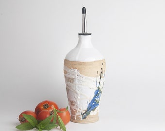 May include: A white ceramic oil and vinegar bottle with a brown, blue, and white glaze. The bottle has a stainless steel pour spout and is sitting on a white surface with tomatoes and green herbs.