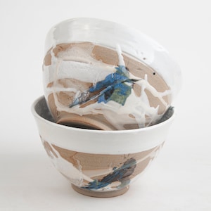 May include: Two ceramic bowls with a white glaze and brown, blue, and green abstract designs. The bowls are stacked on top of each other.