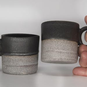 May include: Two ceramic mugs with a black and white glaze. The mugs have a rounded shape and a handle.