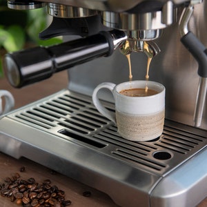 May include: A stainless steel espresso machine with a white ceramic mug on the drip tray. The machine is dispensing coffee into the mug.
