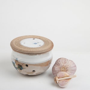 May include: A ceramic garlic keeper with a lid, in white and tan with blue floral details. Two garlic bulbs are next to the keeper. The keeper has small holes for ventilation. The keeper is approximately 10 cm in diameter.
