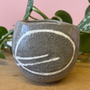 Puede incluir: Una pequeña taza de cerámica redondeada con un acabado texturizado, gris oscuro y moteado. El exterior presenta un diseño de línea circular blanca pintado a mano que envuelve el lateral. El borde es ligeramente irregular y la superficie tiene un aspecto mate y terroso.