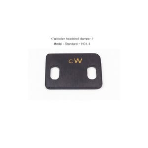 Peut inclure: Un amortisseur de tête de lecture en bois noir avec deux trous circulaires et les lettres dorées "cW". Le texte au-dessus indique "Wooden headshell damper" et "Model: Standard - HD1.4".