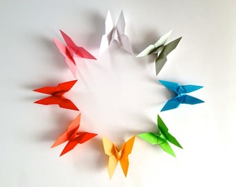 May include: Eight origami butterflies arranged in a circle on a white background. The butterflies are in a rainbow of colours, starting with pink and ending with orange.