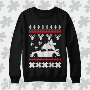 Op de afbeelding: Zwarte kersttrui met een wit silhouet van een auto die een kerstboom draagt. De trui heeft een rood en wit sneeuwvlokkenpatroon.