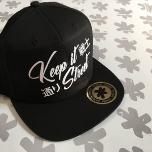 Op de afbeelding: Zwarte baseballpet met een wit geborduurd logo met de tekst "Keep it Street" in een gestileerd lettertype. Het logo bevat ook Japanse karakters.